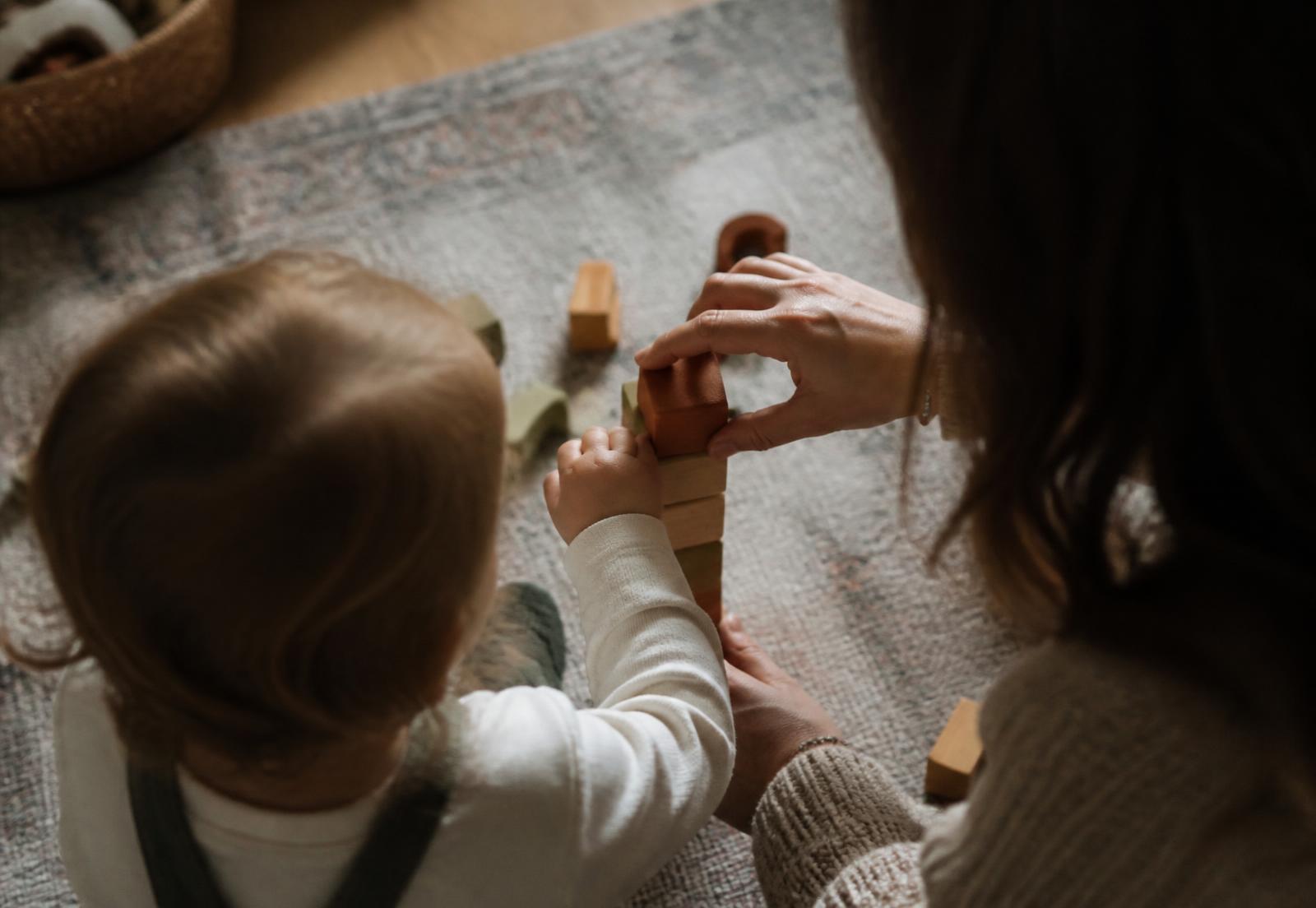 A parent and a toddler stacking wooden blocks together on a rug at home