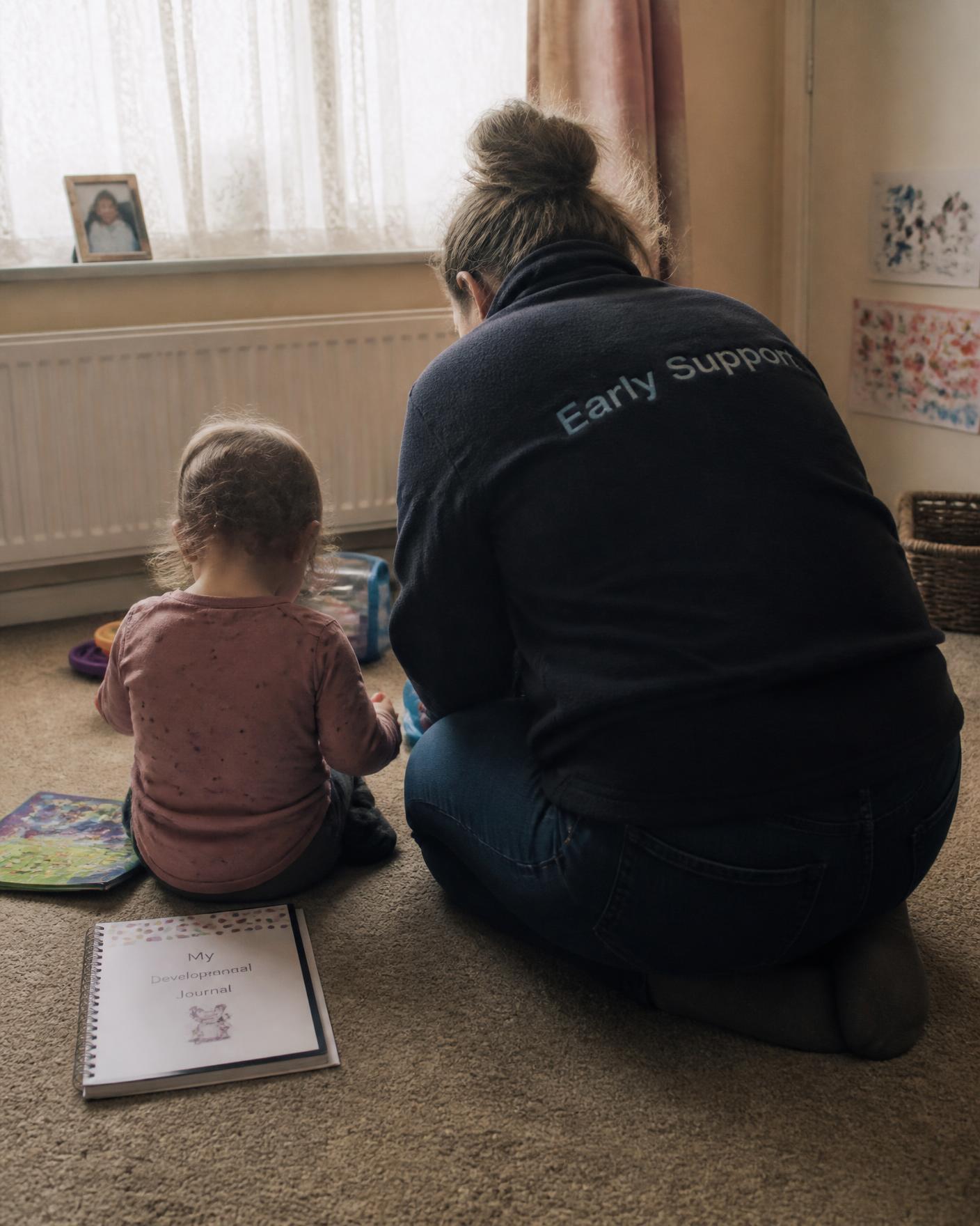 A Portage home visitor kneeling beside a young child on a living room carpet, both seen from behind, with a developmental journal on the floor