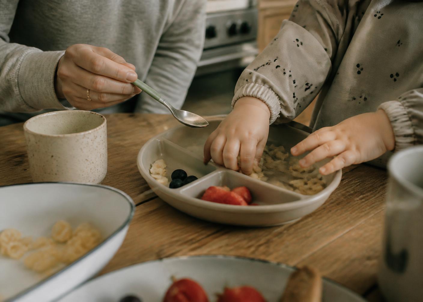 A toddler's hands exploring food on a divided plate while a parent offers a spoon