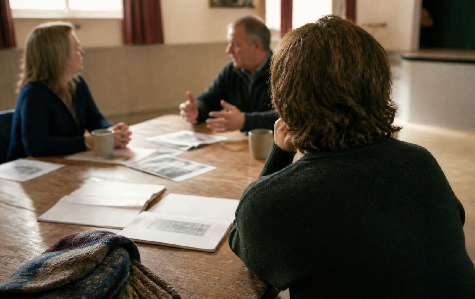 Three adults talking around a wooden table in a village hall with mugs of tea and papers